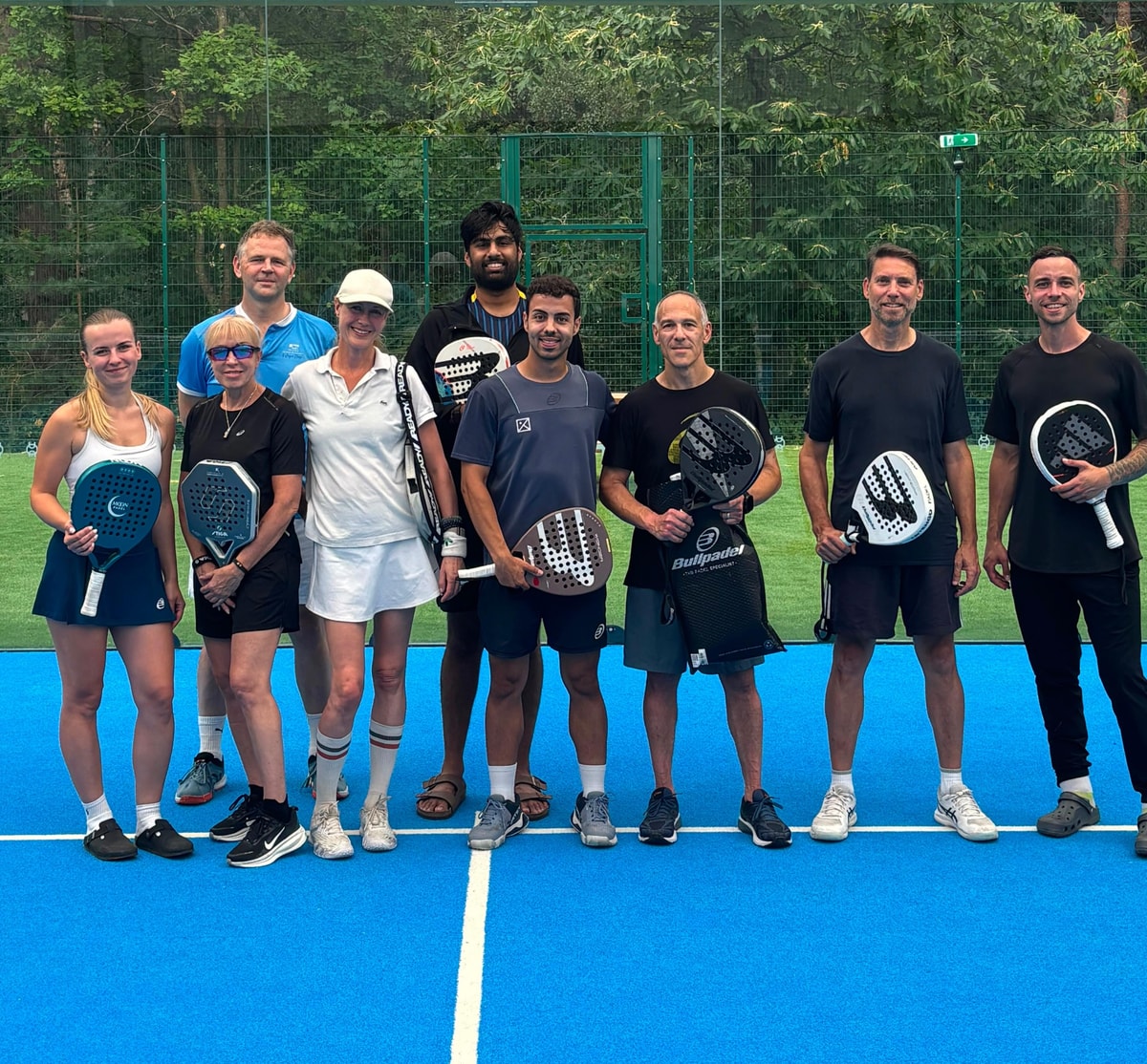 Nine padel players of mixed ages line up with their rackets on a blue court in front of a fence and trees