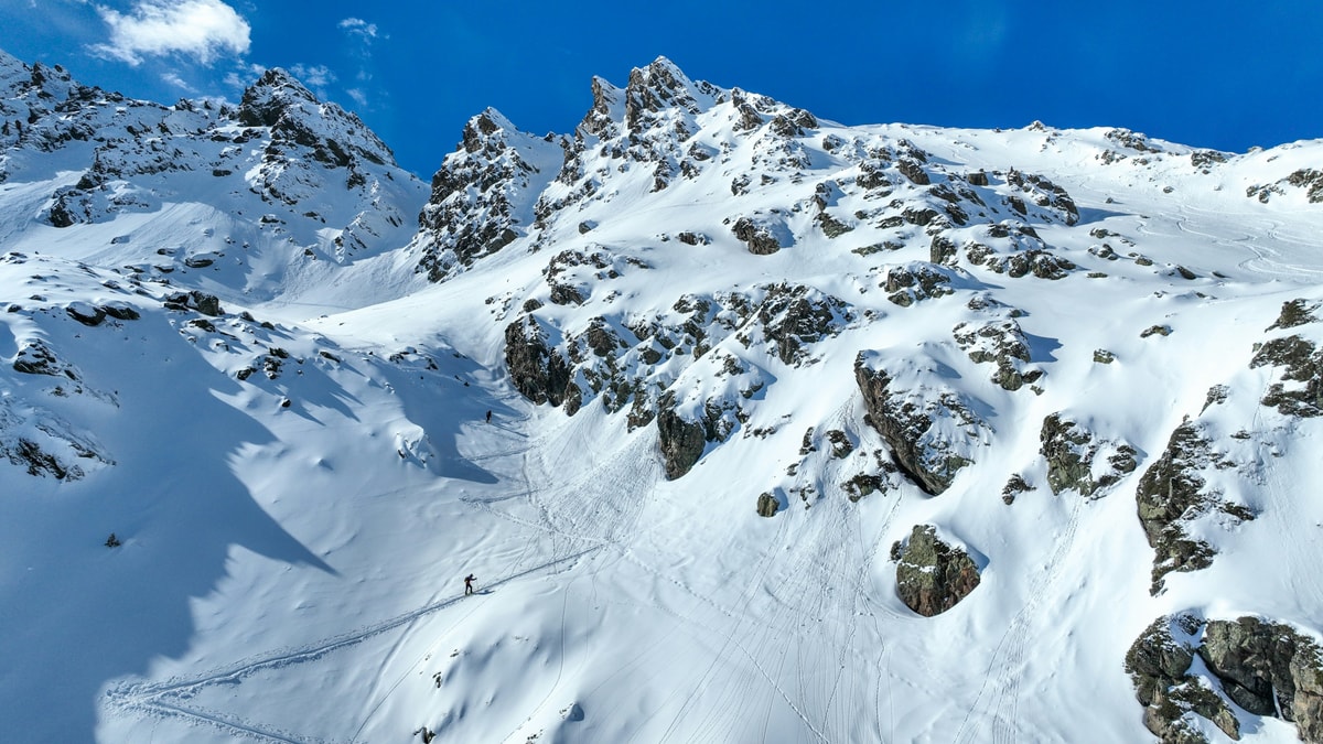 A lone ski tourer climbs a snowy bowl below jagged rocky peaks in Andorra under a deep blue winter sky